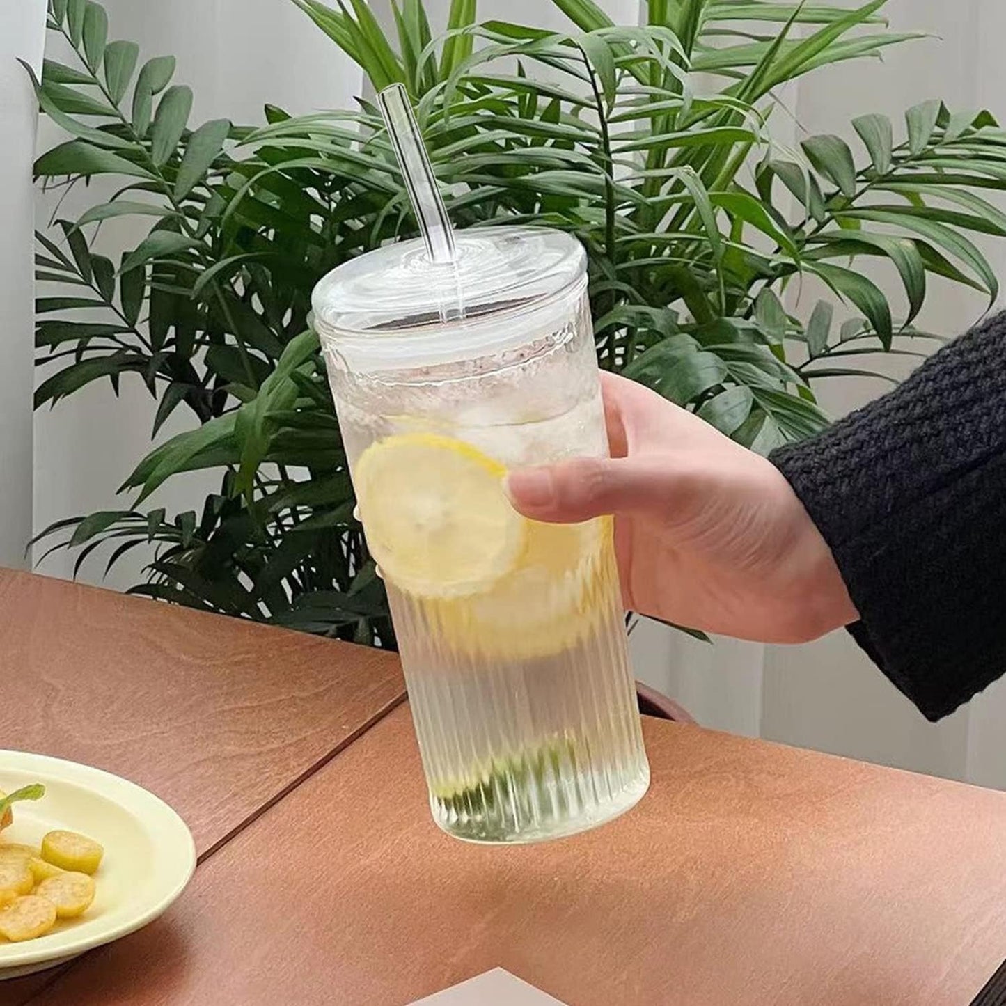 Bir el, erkugo. tarafından yapılmış, borosilikattan yapılmış, kapak ve pipet ile tamamlanmış şık bir Macchiato Sunum Bardağı tutuyor. Berrak sıvı ve limon dilimi ile dolu bardak, atıştırmalıkların yanındaki ahşap bir masanın üzerinde duruyor. Yeşil bitkiler dingin bir arka plan ambiyansı yaratıyor.