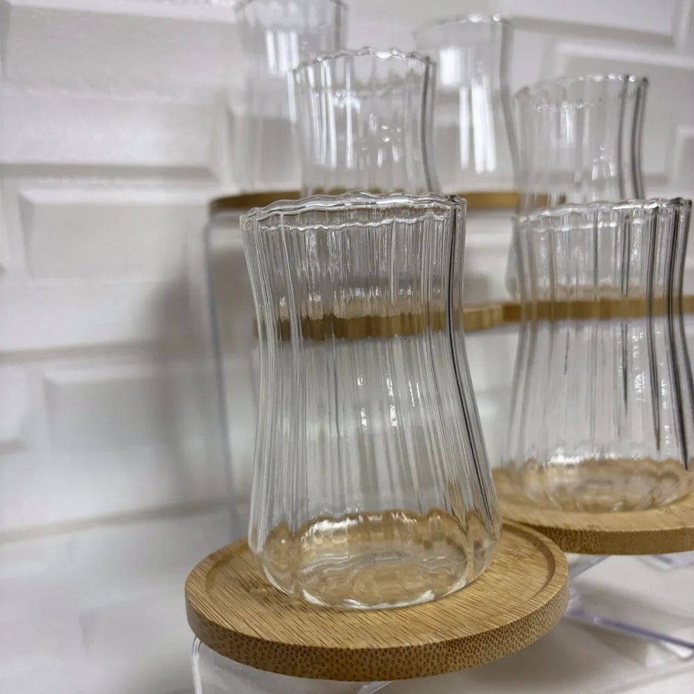 Erkugo'nun Ehlikeyf Çay Bardağı borosilikat cam bardakları, 6'lı set halinde, dokulu beyaz bir duvara karşı ahşap bardak altlıklarında sergileniyor.