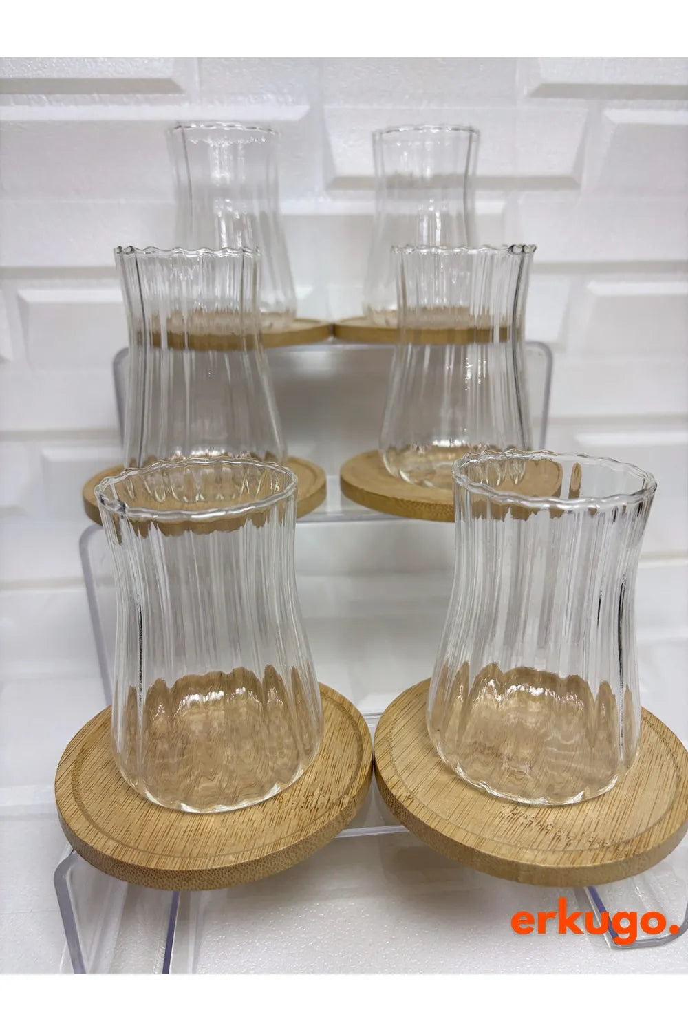 Erkugo'dan Ehlikeyf Çay Bardağı, Borosilikat 6'lı Set. ahşap bardak altlıkları üzerinde altı adet şeffaf, flütlü cam bardaktan oluşur ve dokulu beyaz bir arka plana karşı katmanlı akrilik bir stand üzerinde zarif bir şekilde sergilenir. Sağ altta turuncu renkteki "erkugo." markası yüksek ısı dayanımı sağlar.